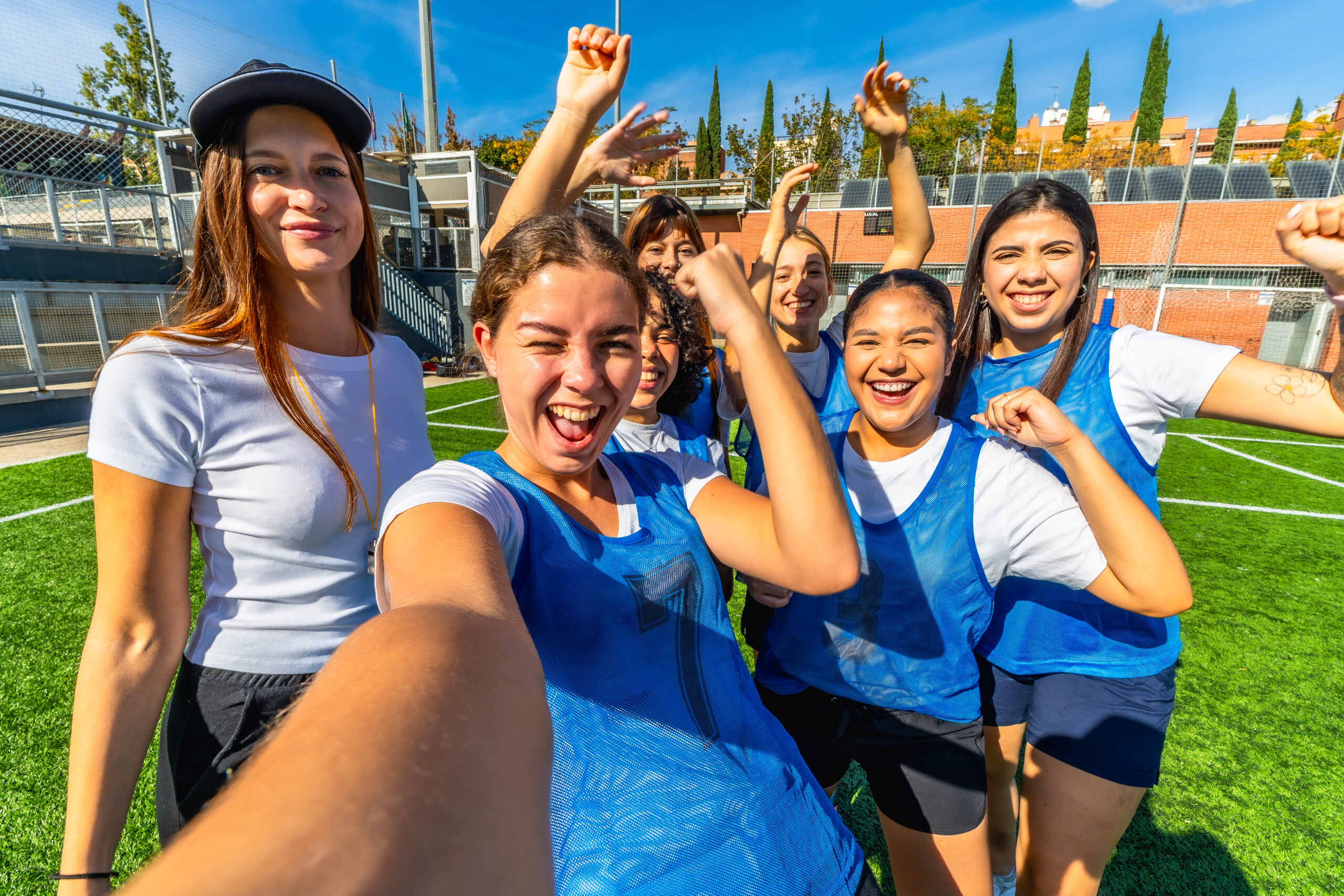 Group of young, multiracial women soccer players cheering and taking a joyful selfie on sunny green turf after a victory, arms raised in celebration and friendship